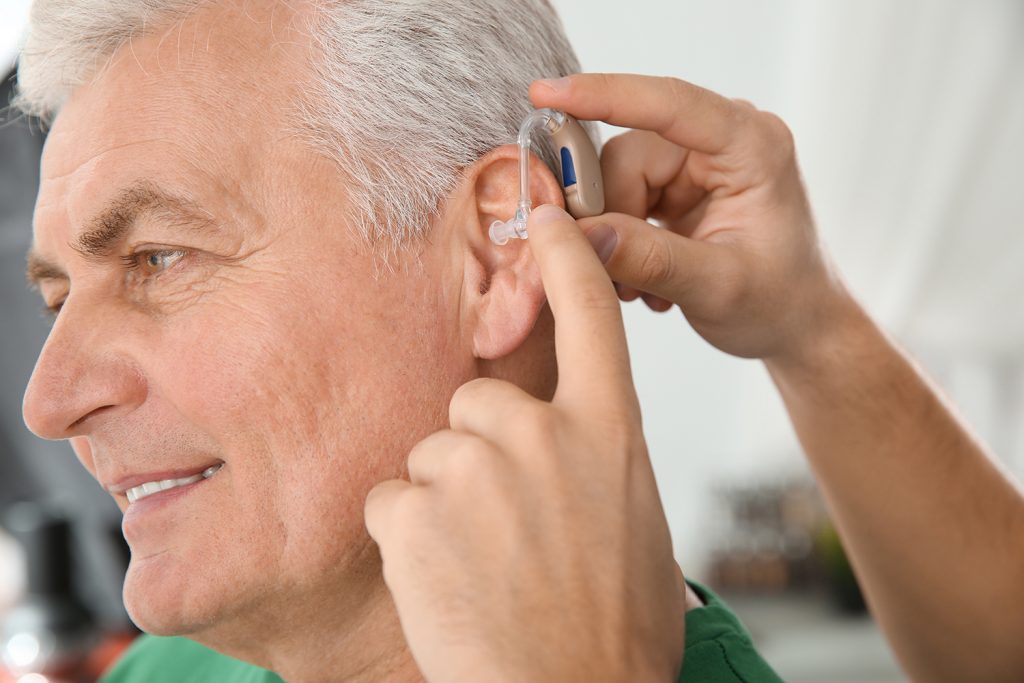 Young man putting hearing aid in father's ear indoors, closeup